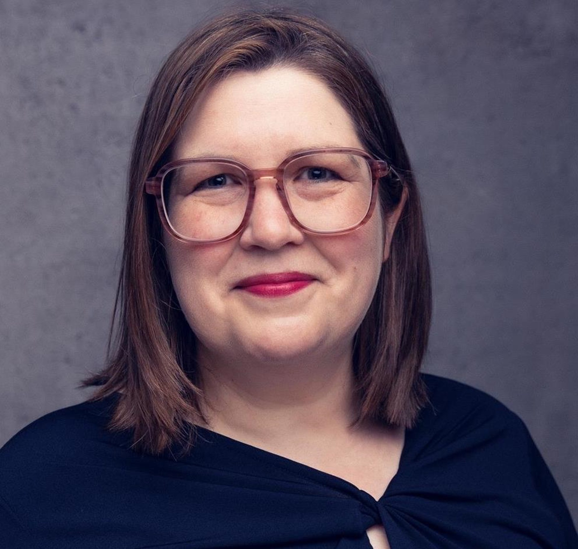 Bei dem Bild handelt es sich um ein Portrait. Eine Frau schaut in die Kamera und lächelt. Sie trägt ein schwarzes Oberteil und eine Brille. 